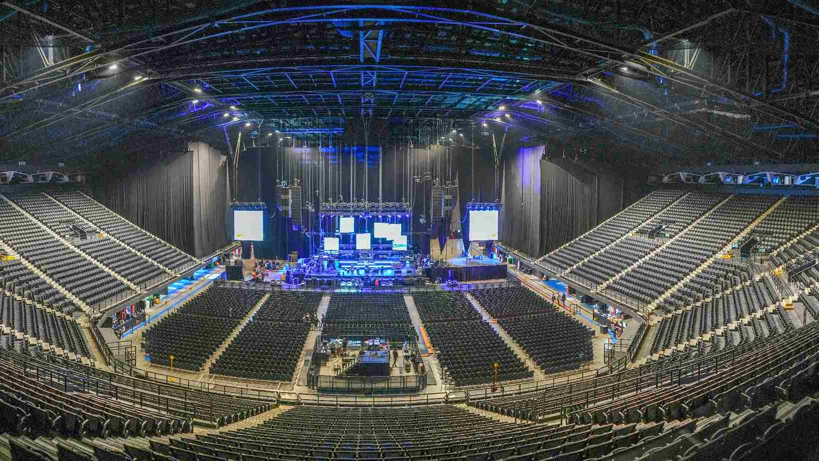 Interior of a large concert venue, empty and with a high ceiling. From above, a stage with giant screens can be seen, surrounded by stands with thousands of seats.