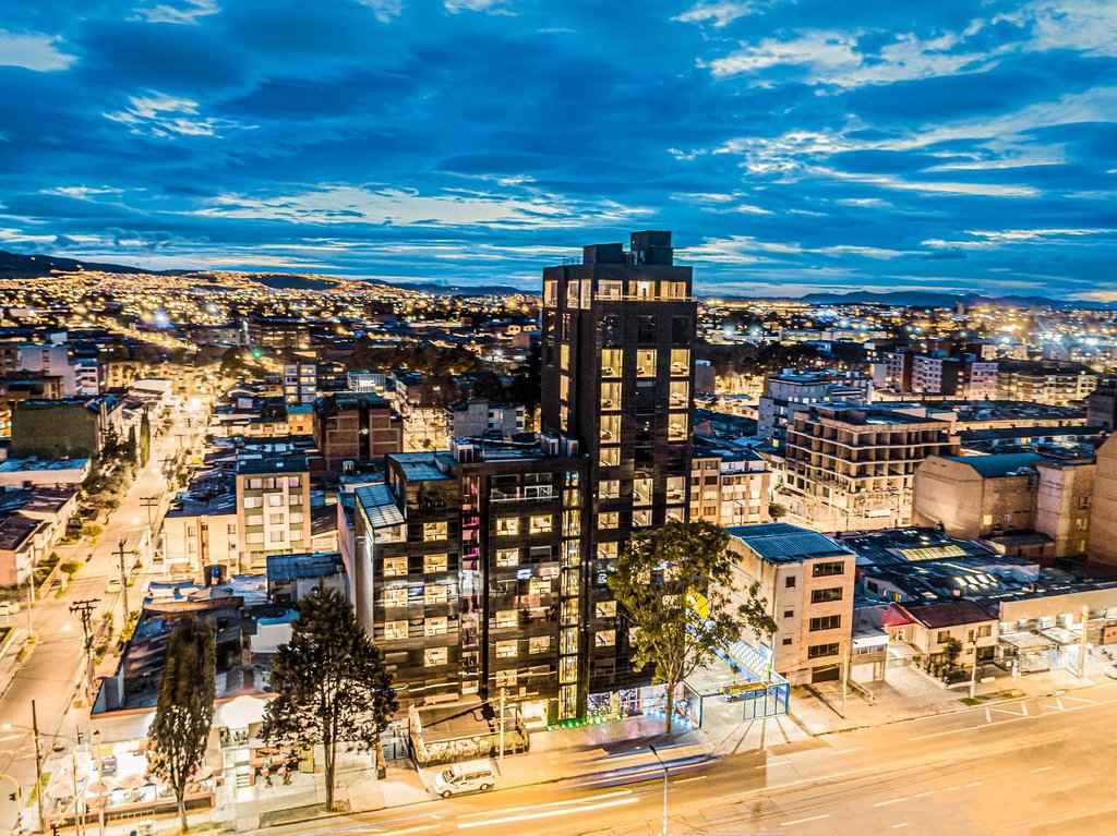 Panorámica al atardecer de un edificio con una alta torre negra al lado de dos más pequeñas. Se observan otras edificaciones iluminadas alrededor. El cielo es muy azul.