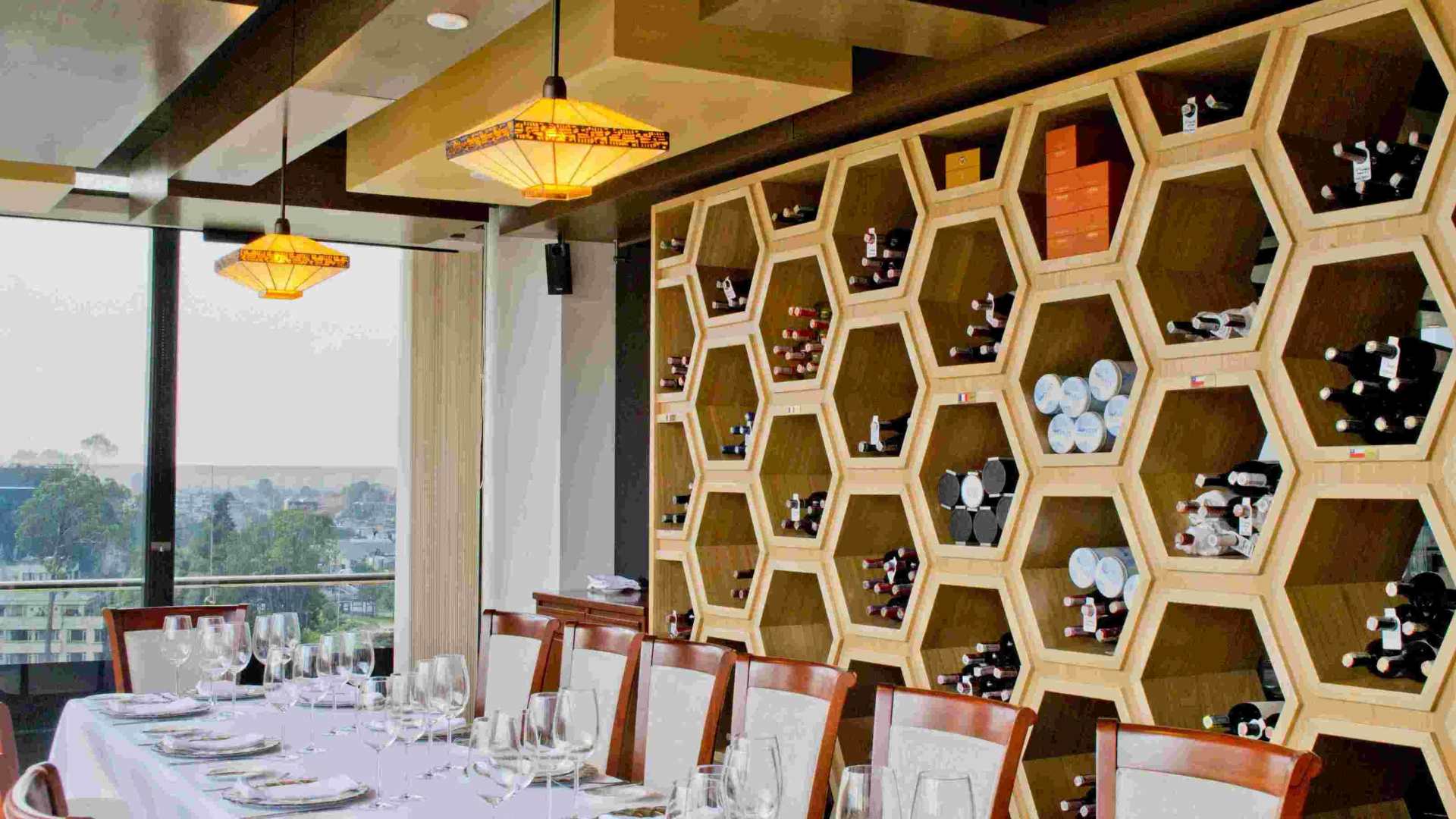 Interior de un salón en el que se observa una elegante cava llena, detrás de una larga mesa decorada con manteles blancos, copas de cristal y platos.