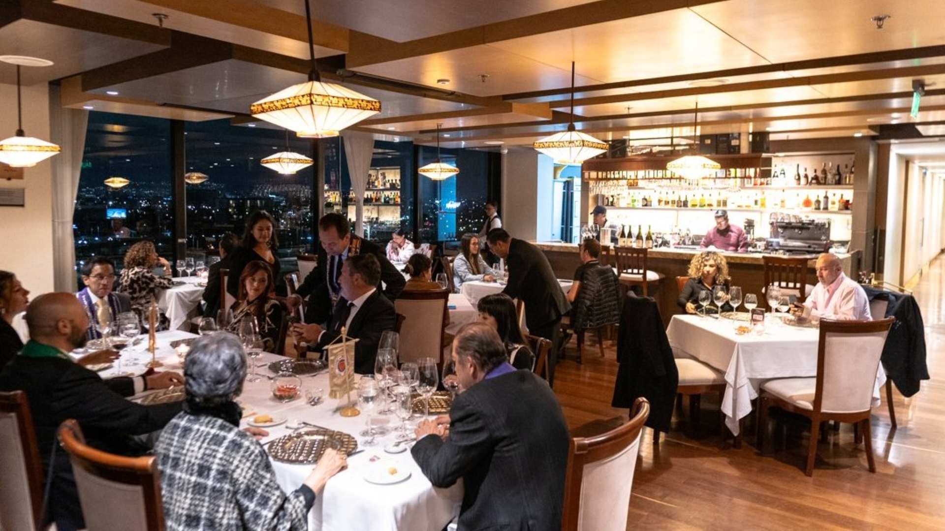  Interior de un salón de noche con varias mesas decoradas con manteles blancos, copas de cristal y platos. Hay varias personas sentadas alrededor de las mesas.
