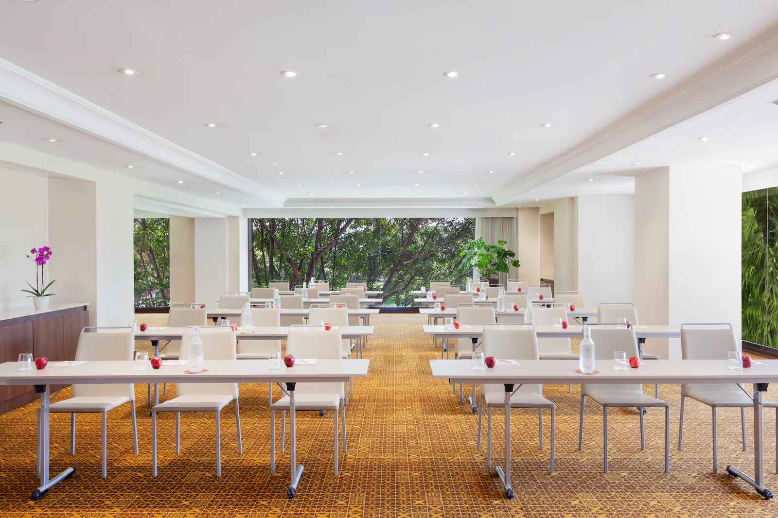 Gran salón con ventanales a jardines a los lados y al fondo. Hay dos filas con 10 de mesas, organizadas de forma vertical. hay un lindo tapete color caoba y una matera con una orquídea.