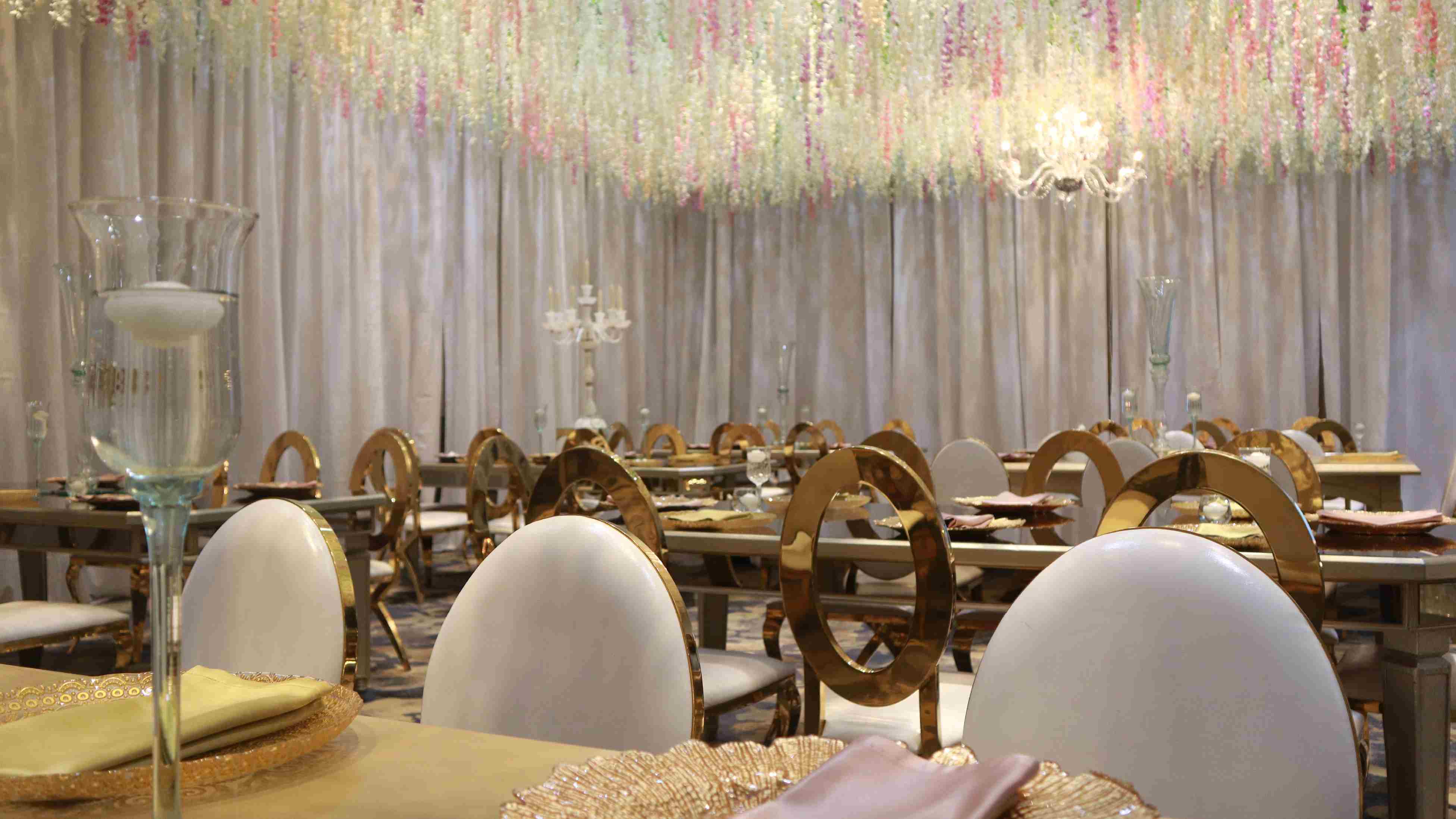 Primer plano de salón decorado elegantemente con mesas y sillas con detalles dorados, un candelabro y un adorno, como una copa larga de vidrio en cada mesa. Las cortinas tienen un detalle decorativo en hilo brillante.
