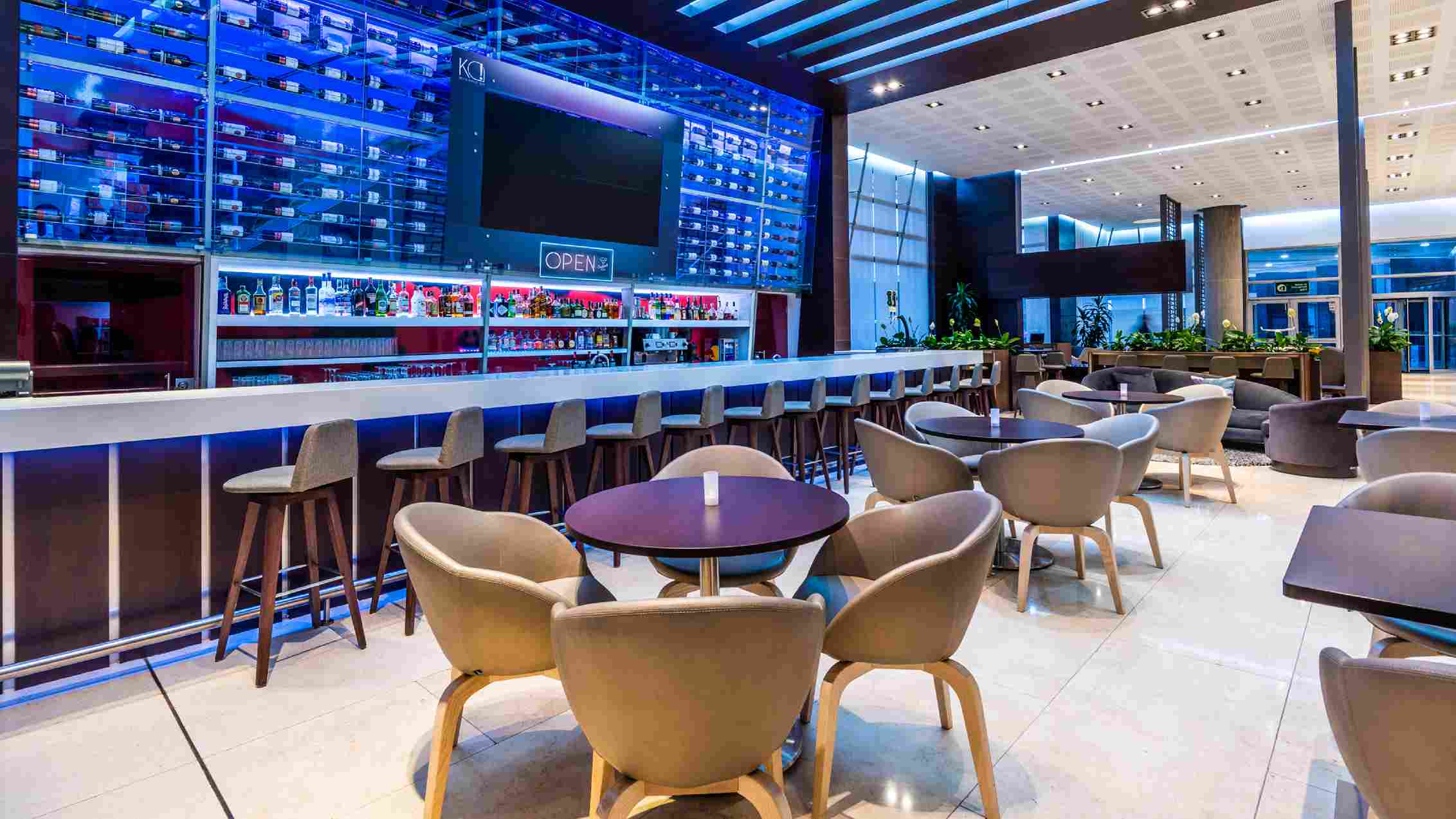  Salón tipo bar grande en el que destaca una barra, una cava en vidrio y varias mesas redondas en madera con sillas alrededor.