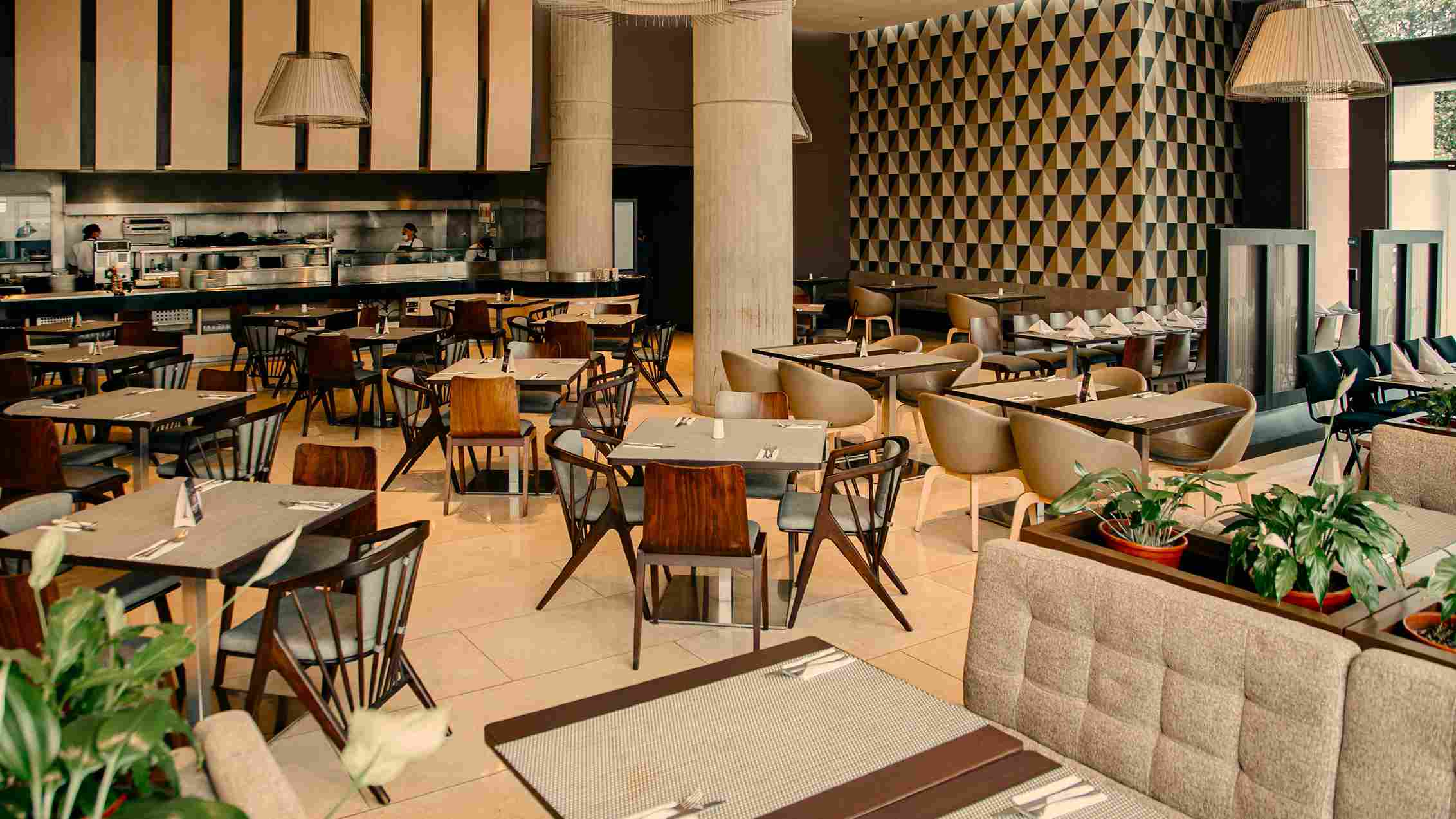  Salón tipo comedor en el que destacan varias mesas con sillas alrededor. Al fondo se observan dos personas en un punto de entrega.de alimentos.