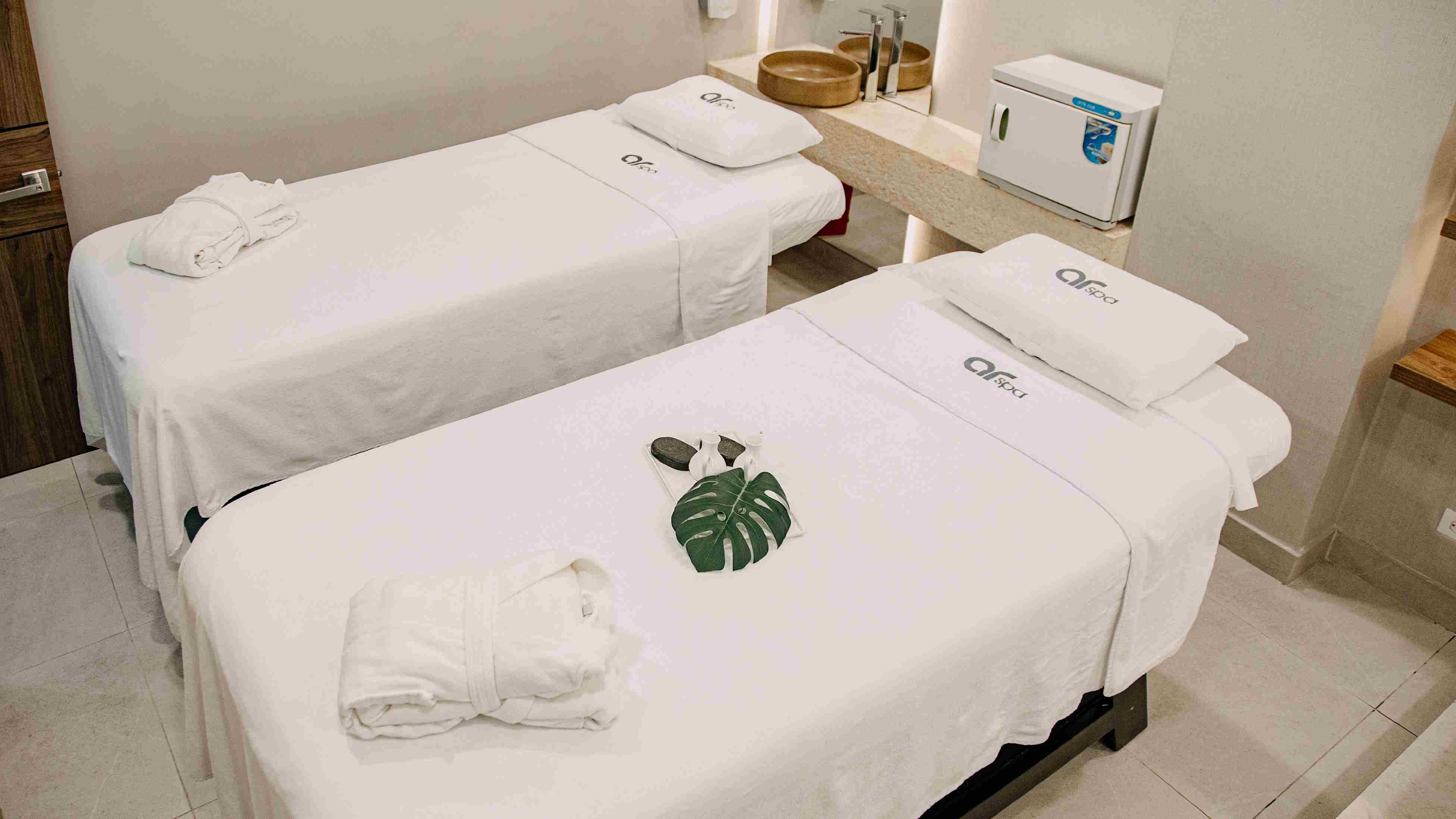 Salón pequeño con dos camas tipo camillas, cubiertas con mantas blancas, Hay dos especies de batas blancas sobre cada cama. En las mantas destaca una leyenda que dice: "ar spa"