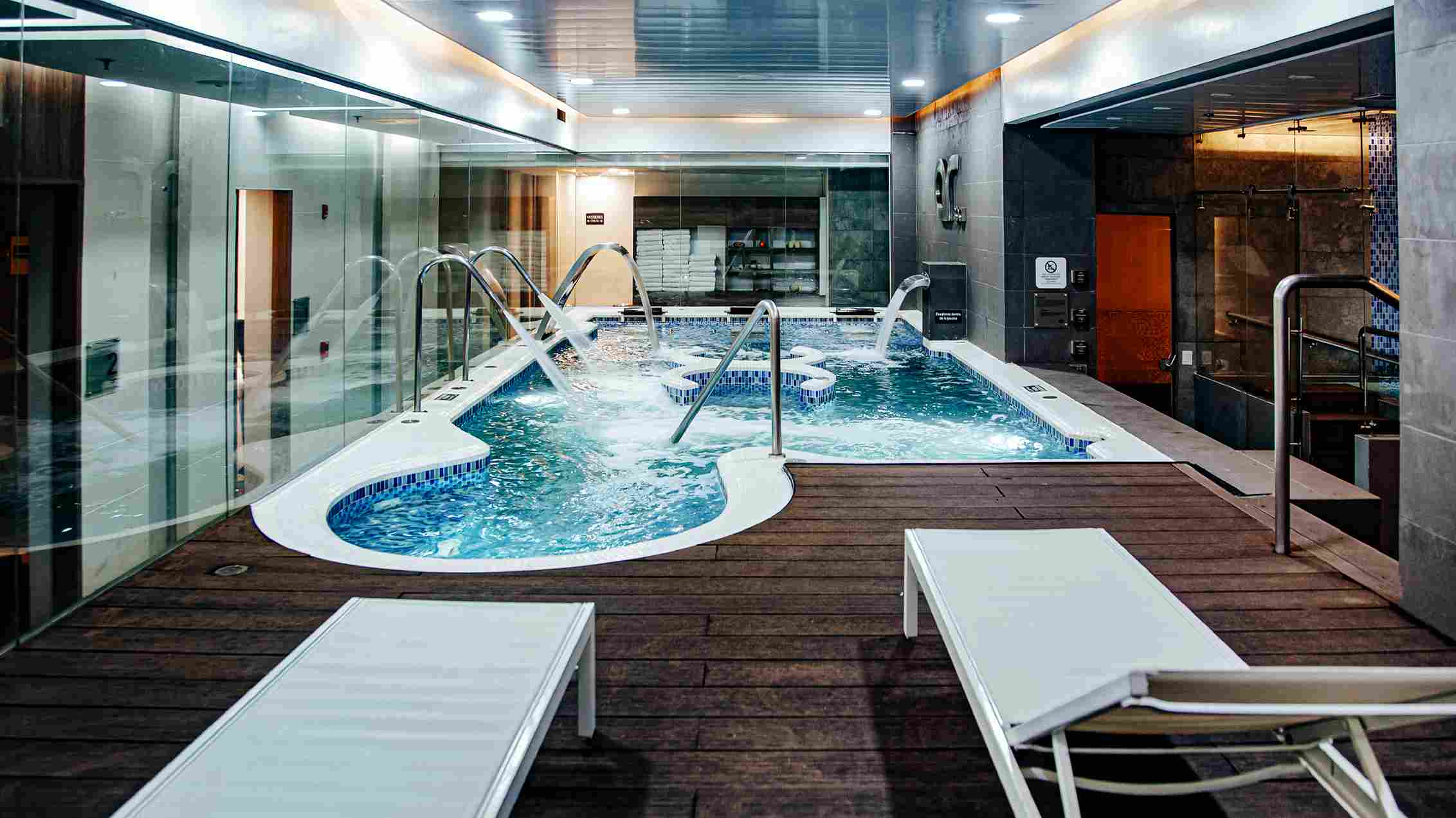  Salón con una piscina en la que las aguas se mueven, generando una capa blanca de burbujas. En el salón que está rodeado por ventanales hay dos sillas largas reclinables.