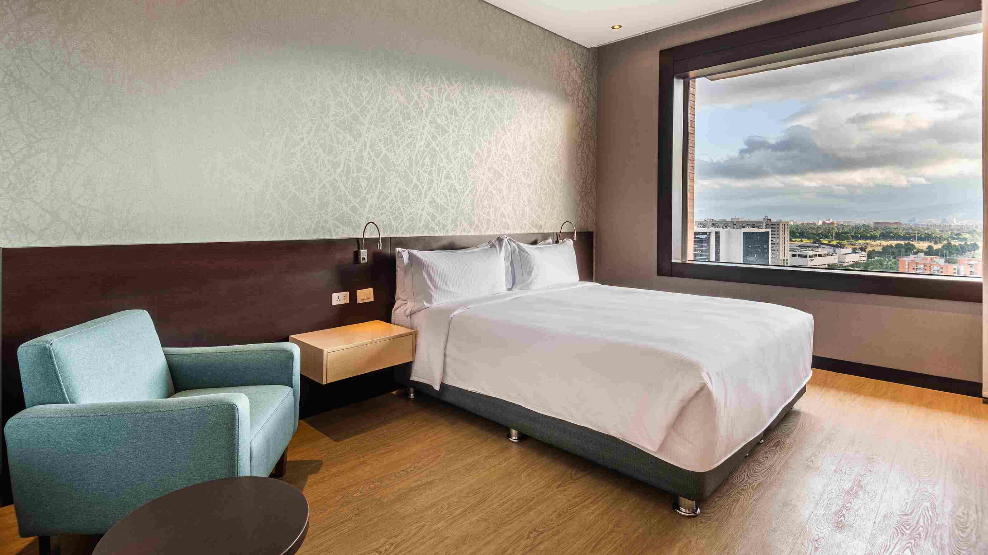 Habitación en un piso alto, con una cama con sábanas blancas y una silla tipo poltrona de color azul celeste. Hay una ventana sin cortinas, desde donde se observa a lo lejos los techos de otras edificaciones.