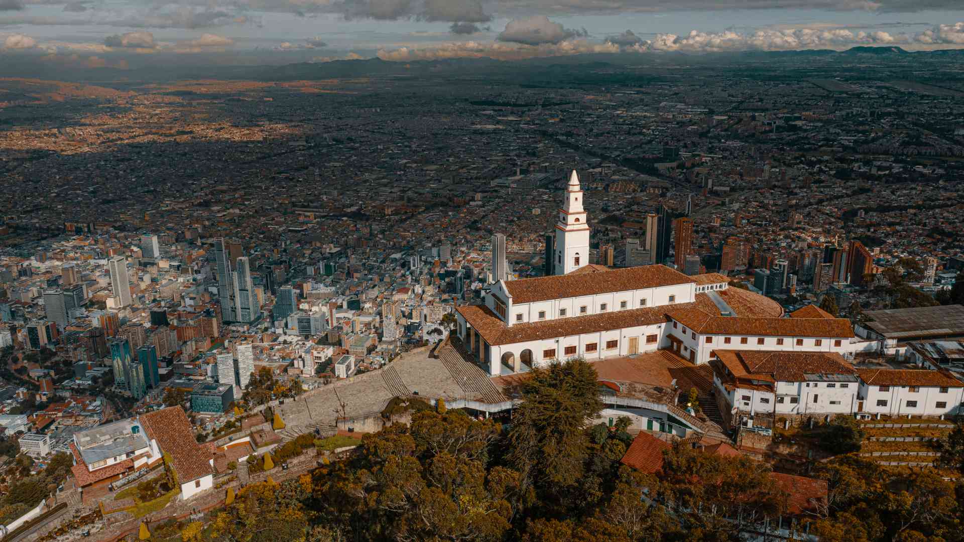 Vista aérea de Monserrate en Bogotá, con su iglesia blanca destacando sobre un vasto paisaje urbano y montañas. 
