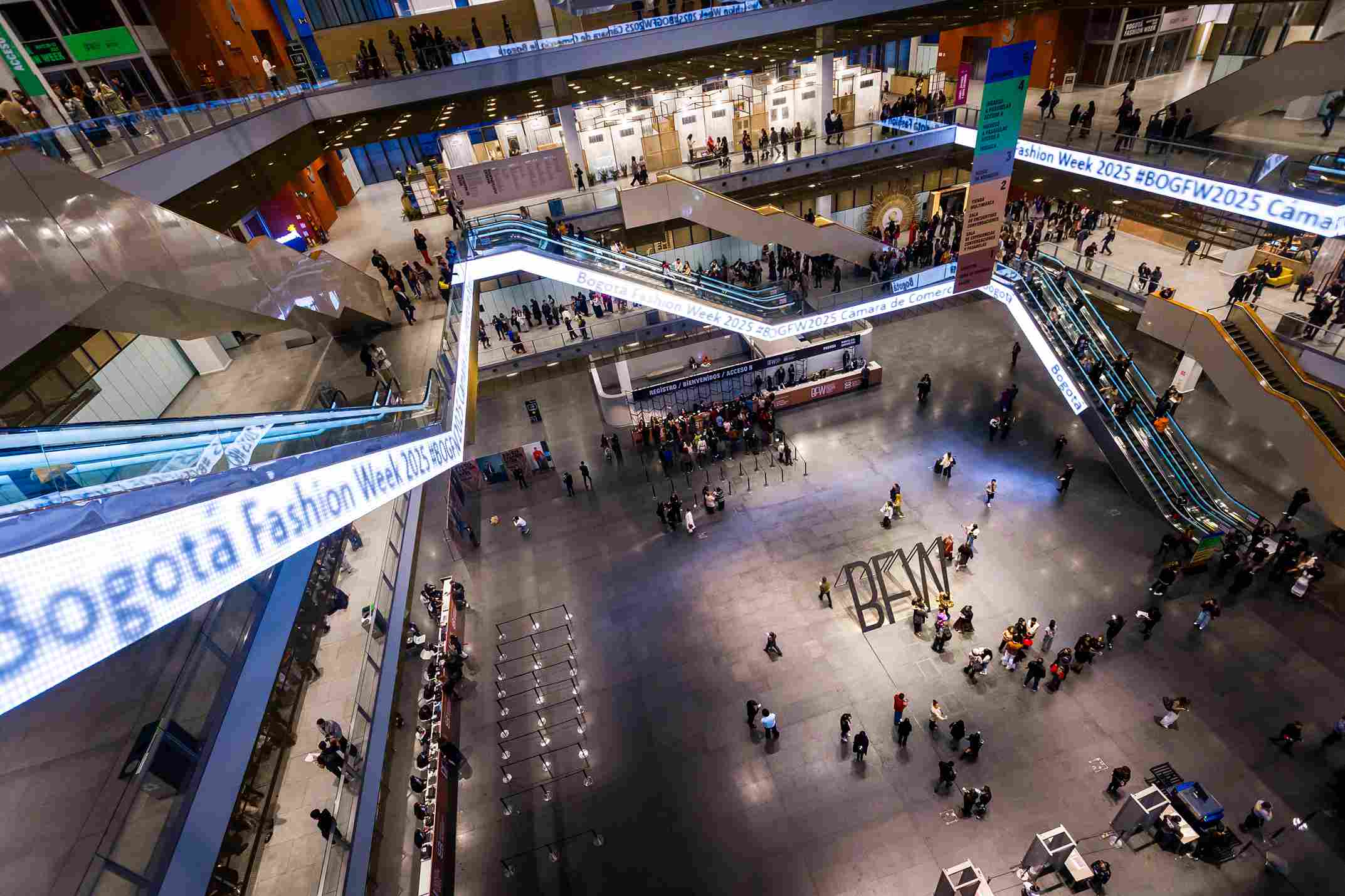 A large indoor business center with people walking and exploring shops and common areas. The view from above reveals the scale of the space and the connections between floors through escalators adorned with striking neon signage.
