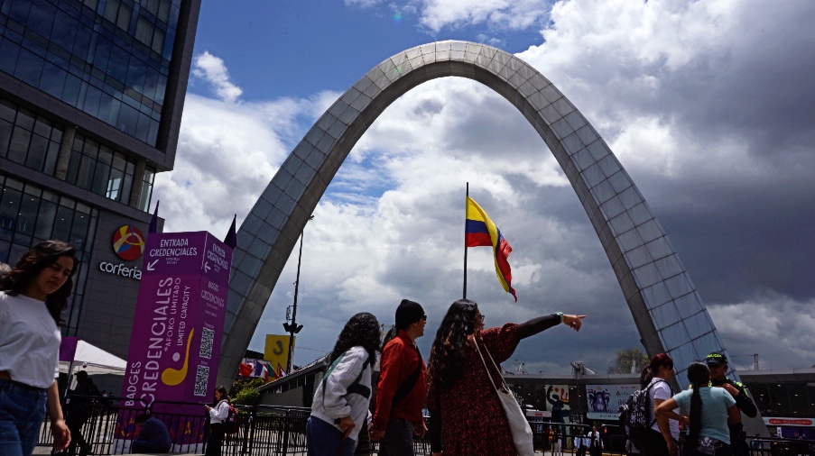 People walk in front of a large arch decorated with a Colombian flag waving on a sunny day.
