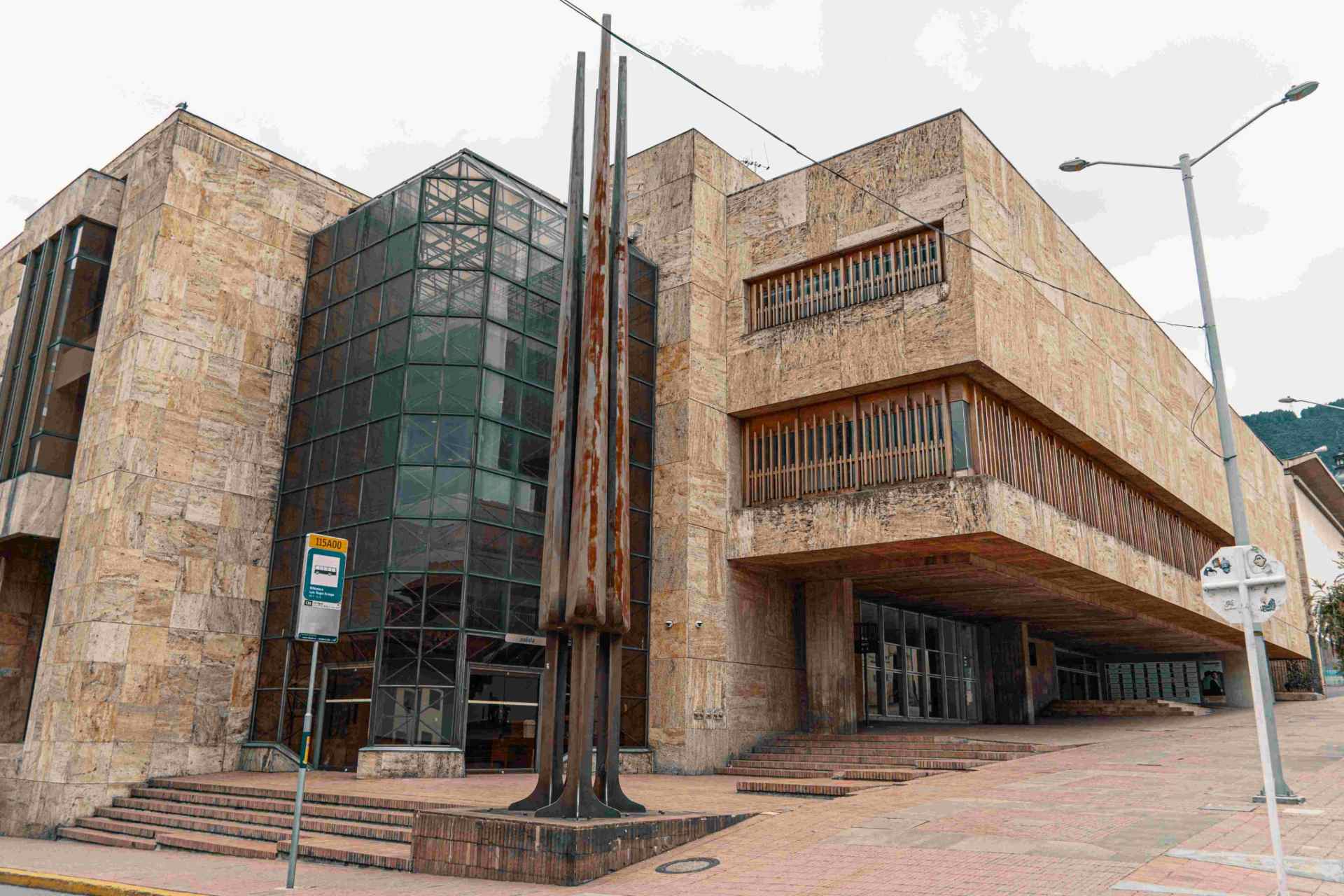 Fachada Biblioteca Luis Ángel Arango de día en ladrillo, baldosa café y vidrio. Monumento metálico al frente.