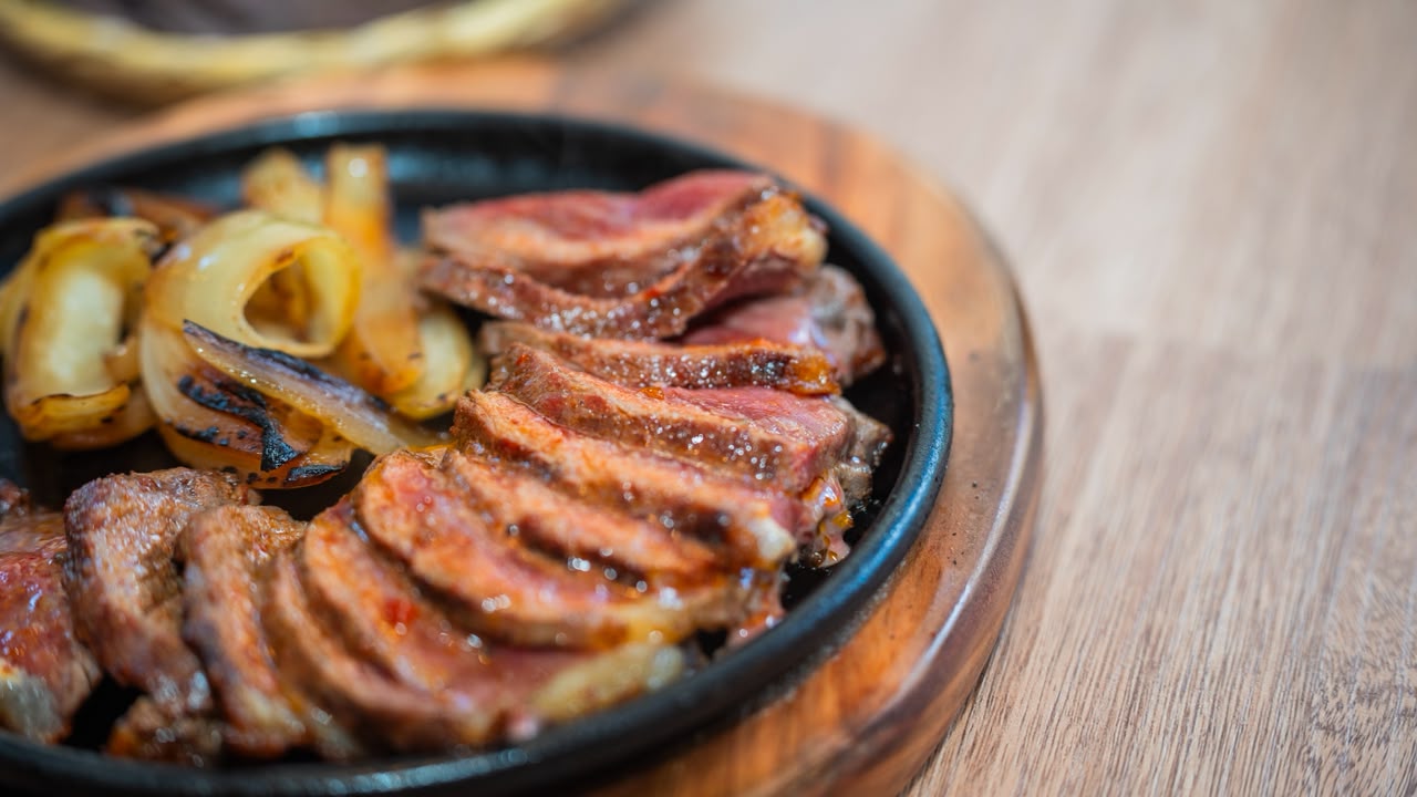 Un plato de carne a la parrilla, cortada en jugosas porciones y servida junto a cebollas asadas. La preparación se presenta en una bandeja de hierro sobre una tabla de madera, en una composición que resalta la textura y el dorado de los alimentos.
