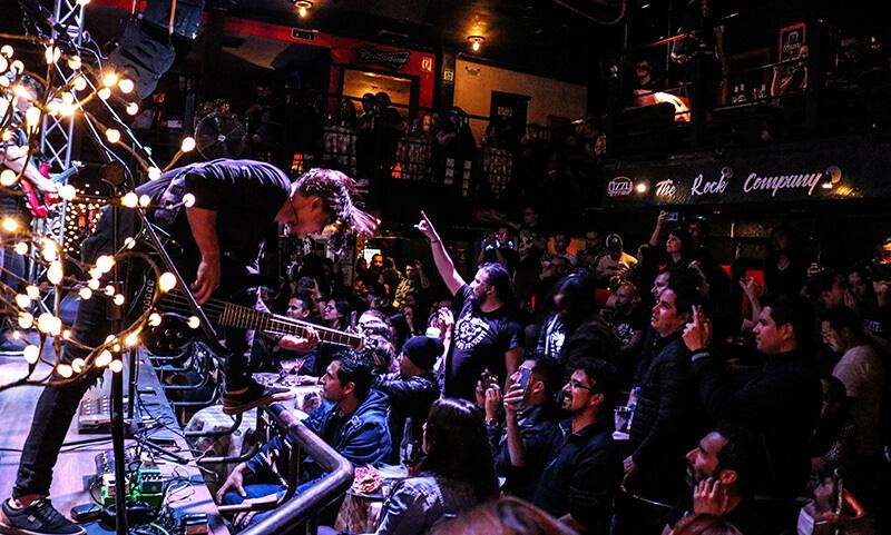 Público de un concierto de rock en un escenario cerrado, con músicos tocando en vivo y asistentes disfrutando de la presentación.