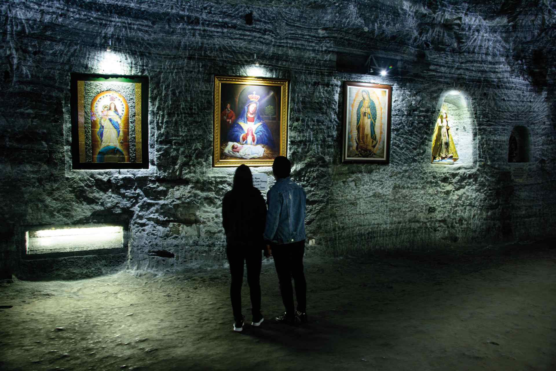 Caverna de sal gris con cuatro cuadros de la Vírgen María. Una pareja de mujer y hombre observan los cuadros.