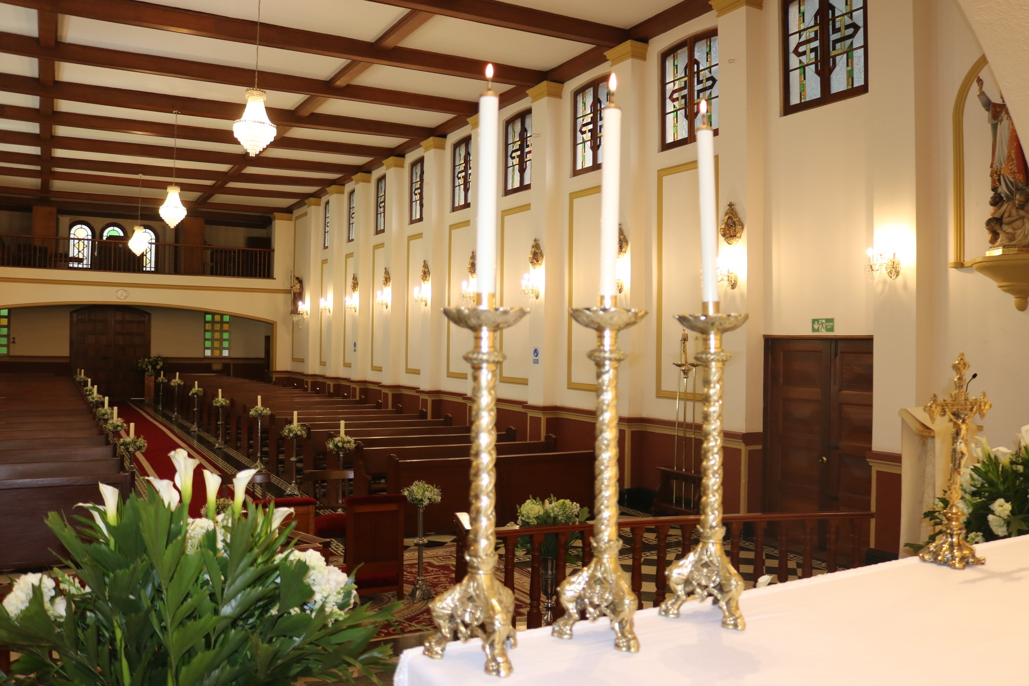 Capilla Liceo de Cervantes interior iluminada con velas y lámparas que cuelgan del techo. Hay arreglos florales al lado de tres velones encendidos sobre el altar. Las paredes son rosadas con rayas doradas y una puerta de madera café. Se ve una fila de por lo menos 20 sillas de culto vacías. Al fondo se alcanza a ver el segundo piso. 