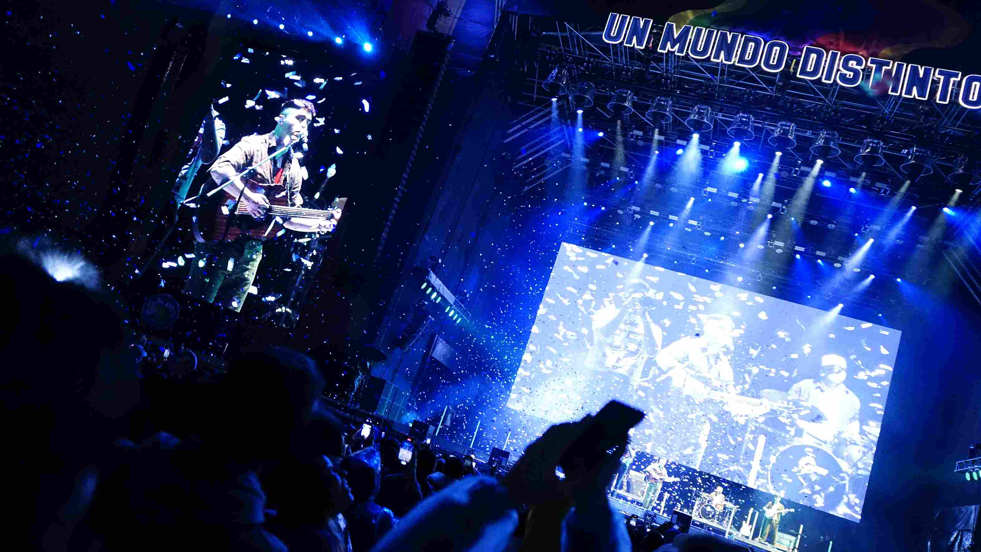 Músico en el escenario con los brazos en alto haciendo la señal del rock, luces de fondo y batería a la derecha.