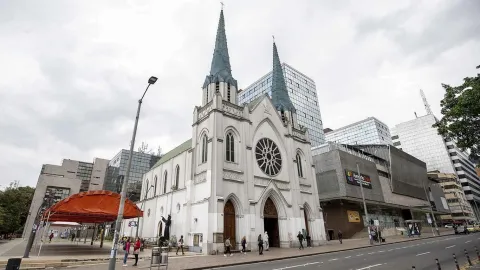 La Porciúncula Church, Gothic-style white façade on Calle 72, with modern buildings and an event tent in front. Several people are walking around. The two gray spires on the roof stand out against the white sky.