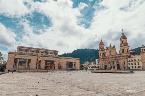 Bolívar Square during the day, seen from the southwest corner looking north, with small pigeons in the distance. In front stands the Palace of Justice, to the side the Primatial Cathedral, and in the background Monserrate Hill under a very cloudy blue sky.