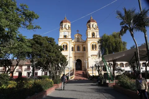 Basílica Menor de San Jacinto fachada de frente vista desde la paza rodeada de árboles y una palma, con un hombre con bastón y una mujer que caminan alejándose de ella. El cielo es muy azul.