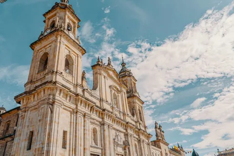 Catedral Primada de Colombia en primer plano contrapicado de la fachada  donde resaltan campanarios y torre del reloj. Se ve el cielo azul con nubes blancas tupidas.