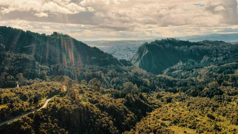 The Eastern Hills with mountains densely covered in trees at sunset, featuring a reflecting rainbow, a desolate winding road, the city in the background, and a cloudy sky.