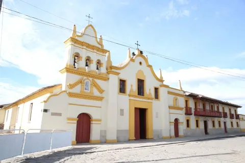Sanctuary of Our Lady of Health – Bojacá, a wide diagonal view of the façade with colonial houses on the left side. The façade is white with yellow stripes, and the bell tower with four bells stands out. The sky is blue with a few clouds.