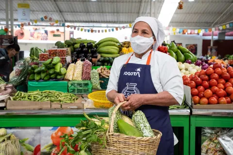 This photograph shows a fruit and vegetable stand inside a market. A woman wearing a chef's hat and white face mask wears a dark blue apron with the "BOGOTÁ" logo. She holds a wicker basket filled with vegetables, including ears of corn and peas. Behind her, a green stall is lavishly stocked with a variety of fresh produce such as cucumbers, eggplants, onions, and large piles of tomatoes. Colorful pennants and lights on the ceiling add a festive touch to the atmosphere.