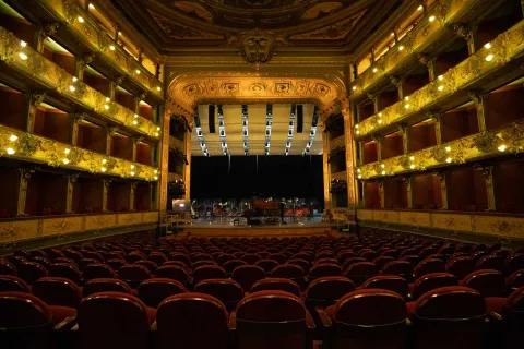 Teatro Colón interior, vista desde la última linea de sillas hasta la tarima donde hay un piano de cola café. En los balcones hay luz encendida.