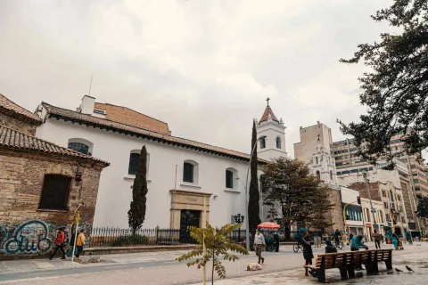 Iglesia de La Veracruz colonial vista lateral con dos pinos y árboles, y al frente la carrera séptima con algunos peatones caminando y otros sentados en sillas de madera. Hay algunas palomas y un perro blanco acostado en la vía.