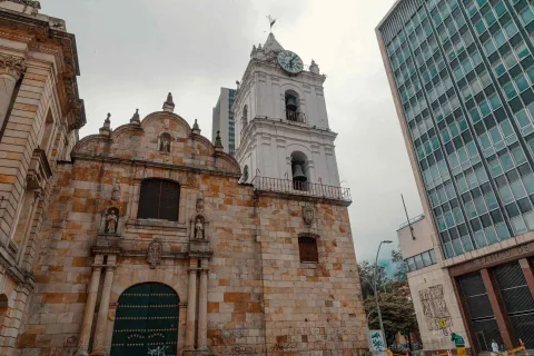Iglesia San Francisco colonial su fachada vista de frente desde la puerta de madera verde y cerrada, hasta la torre blanca del campanario y reloj. Al lado izquierdo se ve la fachada del edificio del Banco de la República.