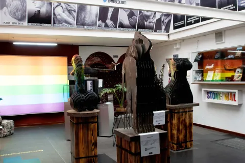 LGBTI Museum, interior exhibition hall with copper artworks, one shaped like a giant phallus and another representing a woman’s body. The room also features photographs of historic gay figures and a screen displaying the colors of the LGBTI rainbow flag.
