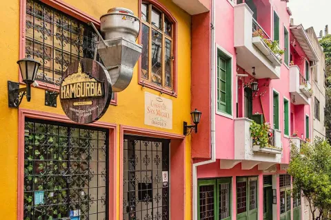 This image of a street in La Macarena captures the vibrant mix of colors that characterizes the neighborhood. In the foreground, a facade in shades of orange, yellow, and red is visible, followed by a pinkish building with white balconies and green windows. The architectural diversity is evident, with a mix of styles and heights giving the neighborhood its eclectic character. The steep streets and the foliage of the trees lining the street are typical features of this bohemian and artistic neighborhood.
