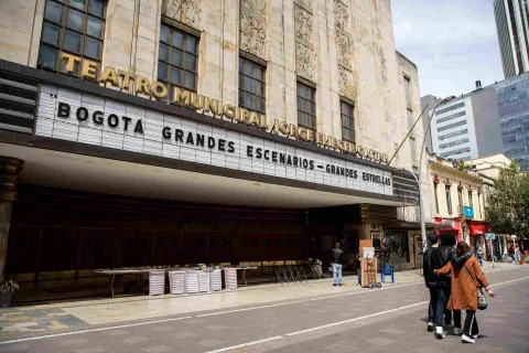 The Jorge Eliecer Gaitán Theater’s facade by day, with its Art Deco doors and walls; the sign bearing the theater’s name in gold capital letters; and the sign announcing the plays on a white background with black capital letters. A few people are walking in front of it.
