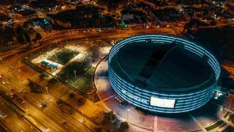 Nighttime panoramic view from above of the Movistar Arena. A couple of synthetic maids can be seen, and in the background some illuminated neighborhood buildings.
