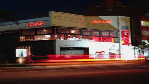 El restaurante "Americana BBQ-Grill Bar" por la noche, con luces de neón rojas y rastros de luz de coches.