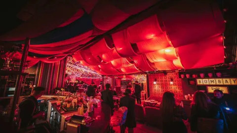 Un bar vibrante y concurrido, "Bombay", con un techo cubierto de telas rojas y luces, creando un ambiente íntimo y festivo.