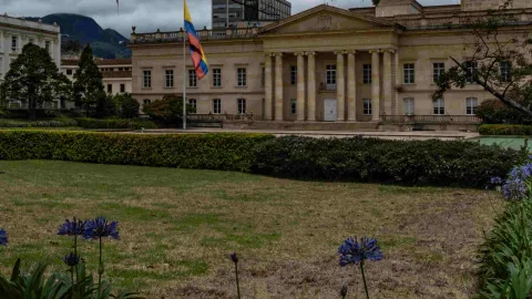 Fachada de la Casa de Nariño, edificio neo colonial en baldoza amarilla en el que se ven cuatro columnas. Al frente hay un amplio jardín con vegetación y una bandera de Colombia gigante.