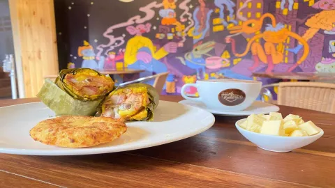 FOTO PORTADA CAFE: Un tamal envuelto en hoja, una arepa de maíz, una taza de café y un tazón de queso, en una mesa de madera.