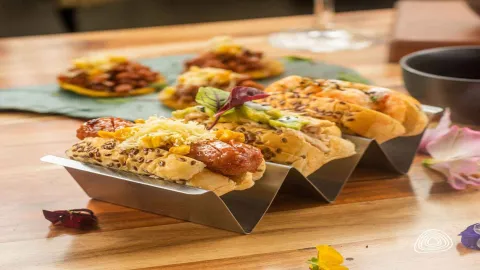 FOTO PORTADA ROCA PAN: Tres mini hot dogs gourmet con aguacate, queso y semillas de sésamo, en un soporte metálico.