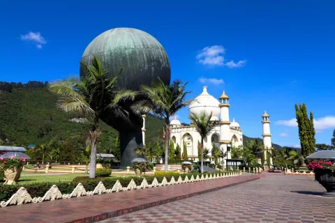 The Hand of God monument, a hand holding a sphere that appears to represent the world, with the Taj Mahal replica in the background. Several palm trees and a mountain in the background complete the setting on a sunny day.