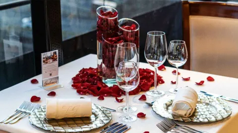 Una mesa elegantemente puesta para una cena romántica, adornada con pétalos de rosa rojos y jarrones altos llenos de rosas.