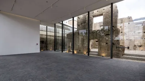 A large, empty room with a large window on the right-hand side, through which a collapsed wall can be seen.