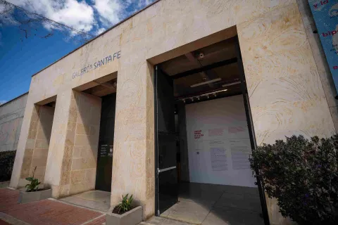 Fachada de edificación en piedra en la que destacan tres puertas negras y altas, parece que de metal, una de ellas abierta y una leyenda sobre la parte superior que dice: “GALERÍA SANTA FE”. Hay tres macetas con plantas pequeñas alrededor de la estructura.