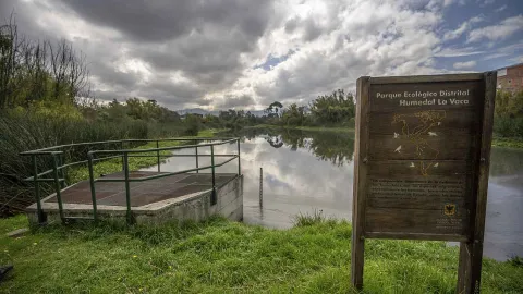 Un espejo de agua en medio de vegetación. Destaca un cartel en madera con una leyenda que dice: “Parque Ecológico Distrital Humedal La Vaca”