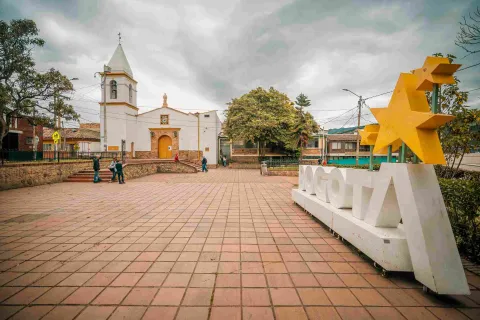 Wide shot of the square featuring a white church with yellow accents in the background. On the right, there is a large white sign with the word "Bogotá", featuring yellow stars above the letter “A.”