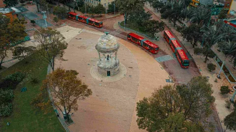 Plaza seen from above, featuring a prominent stone pavilion surrounded by vegetation. A few red TransMilenio buses pass by.