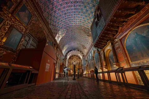 Interior de un recinto religioso, adornado por varios cuadros y retablos. Hay tre hombres que observan el techo y las paredes del lugar.