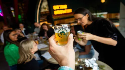 Una mano sostiene un vaso de cerveza artesanal, mientras un grupo de amigos disfruta en el fondo de un bar moderno. 
