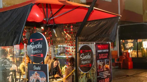 Puesto de comida al aire libre llamado "Sushi Bogotá D.C." con carteles de menú y clientes sentados.