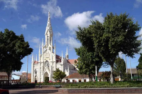 Plano general de la iglesia de estilo gótico con varias puntas hacia el cielo y de color crema. Hay un parque con árboles alrededor.